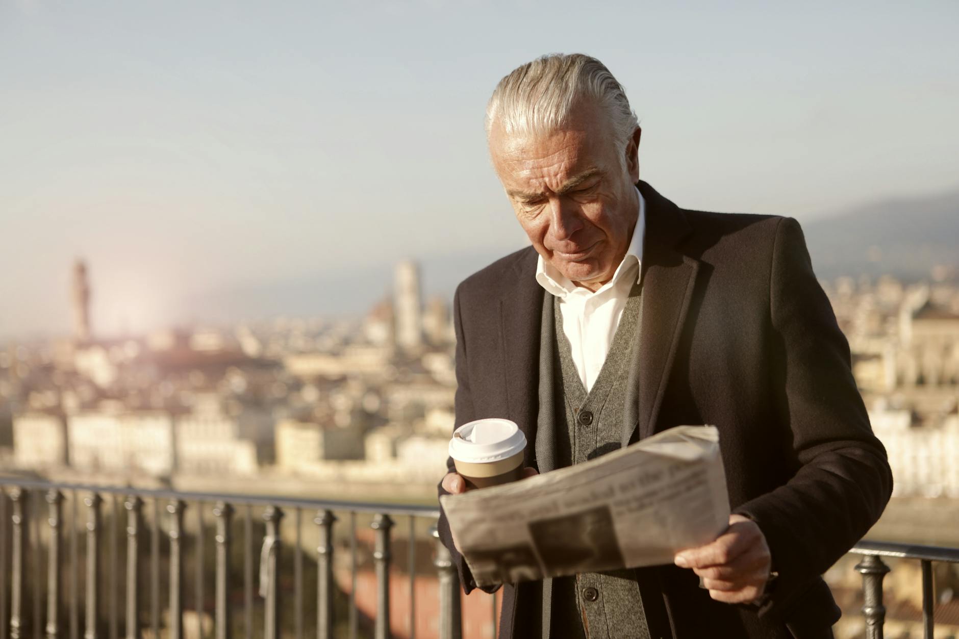 Senior man enjoys coffee while reading a newspaper with a cityscape view.