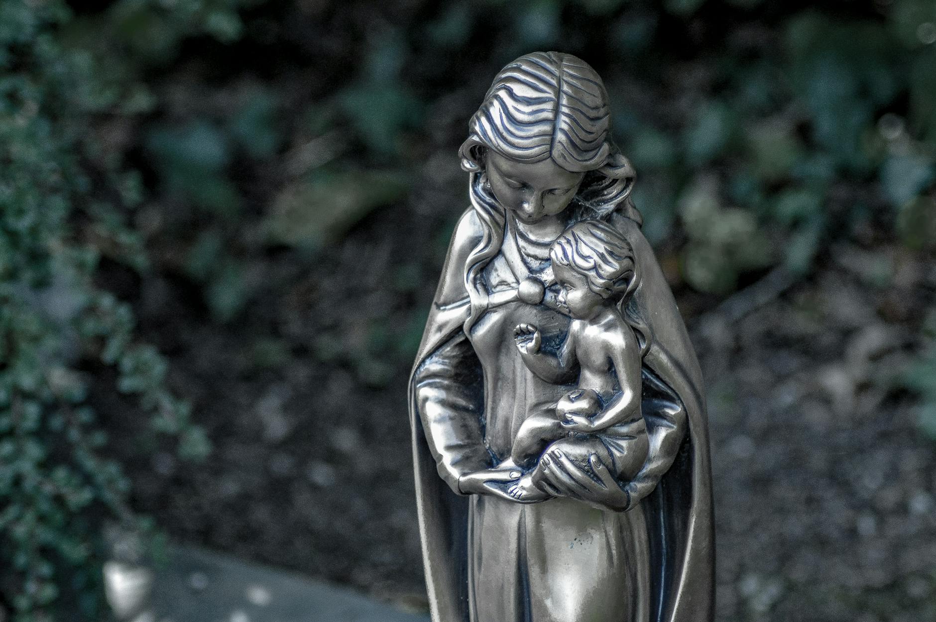 A vintage statue of a mother holding her child, set in a garden, symbolizing maternal love.