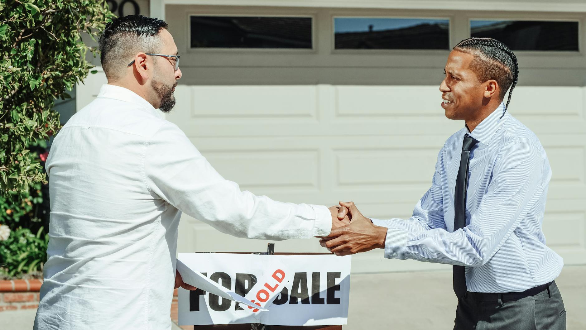 Two men shaking hands in front of a sold house sign, sealing a real estate transaction.
