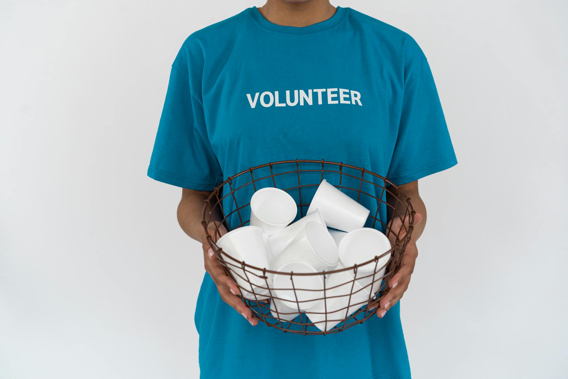 A volunteer in a blue shirt holding a wire basket with disposable cups, promoting recycling.