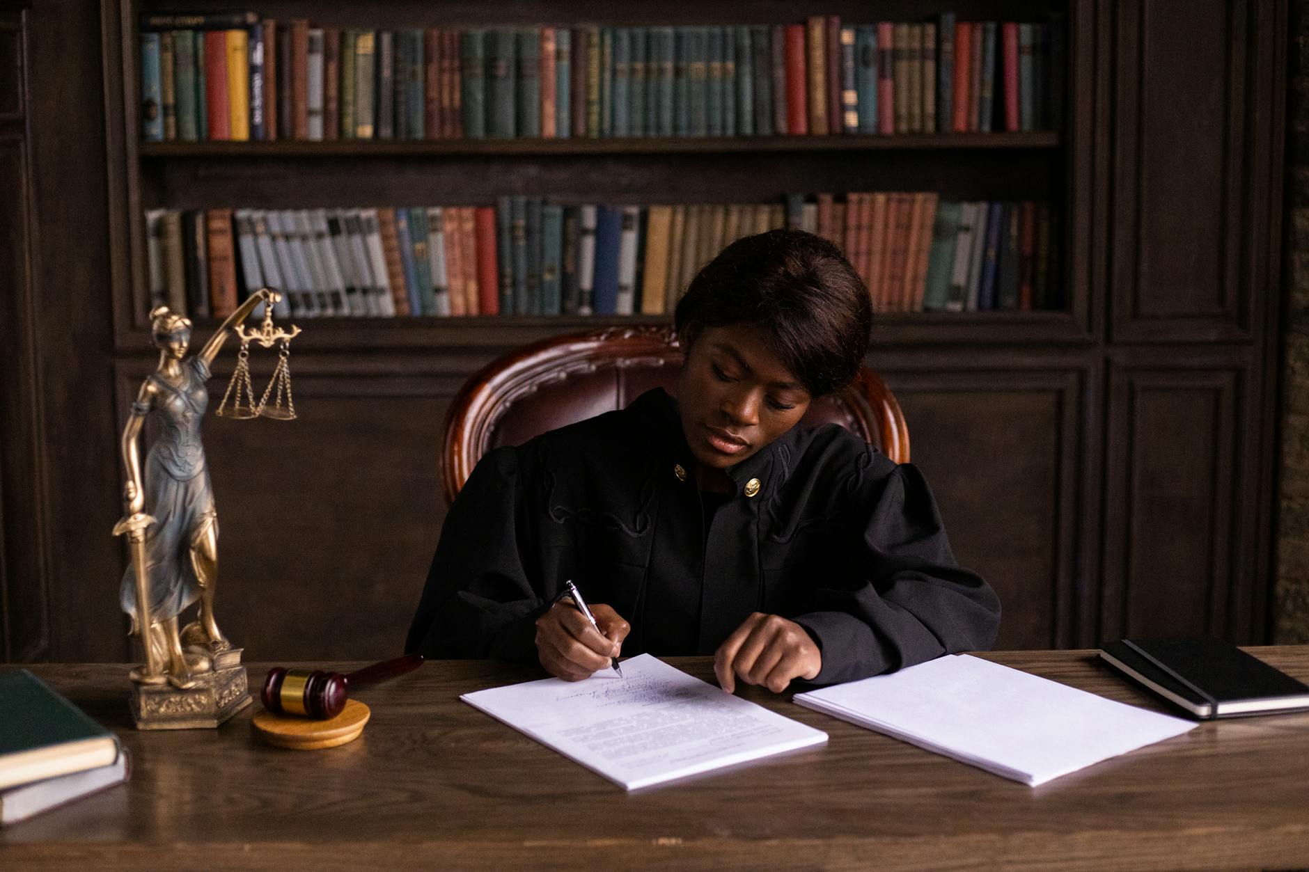 A judge in robes writing on a document at a desk in an office library with law books.